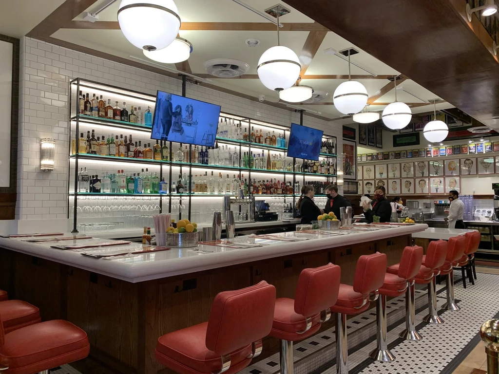 Restaurant bar with illuminated bottles, red stools, and subway tiles.