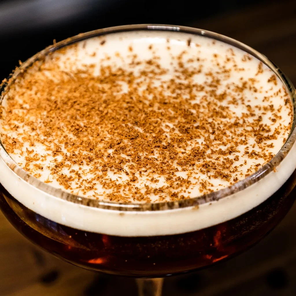 Dark beverage in a stemmed glass, with foam and dark flakes.