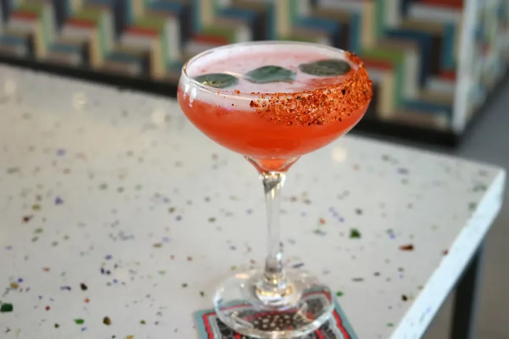Red cocktail in coupe glass with spice rim and green leaves.