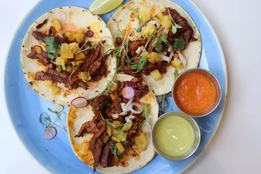 Three tacos with meat, pineapple, cilantro, and radish on a plate.