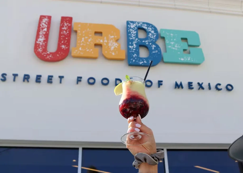 Hand holds layered cocktail with mint and citrus; "URBE" sign.
