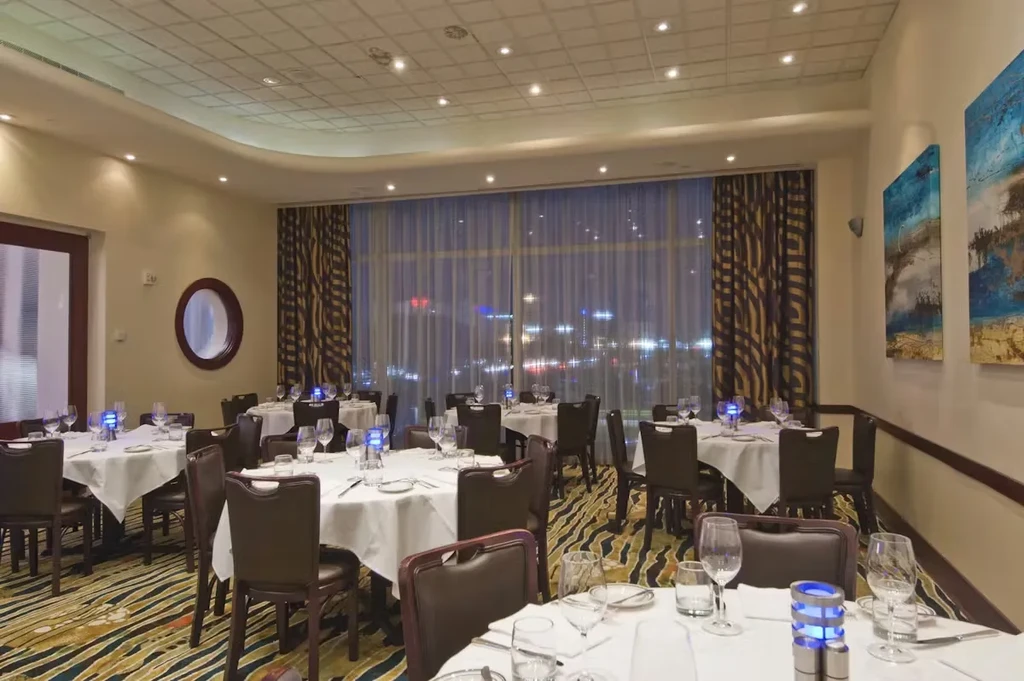 Restaurant dining room with set tables, city lights visible through window.