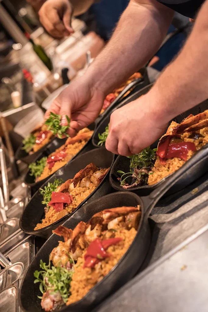 Hands plating seafood paella into dark oval serving dishes.