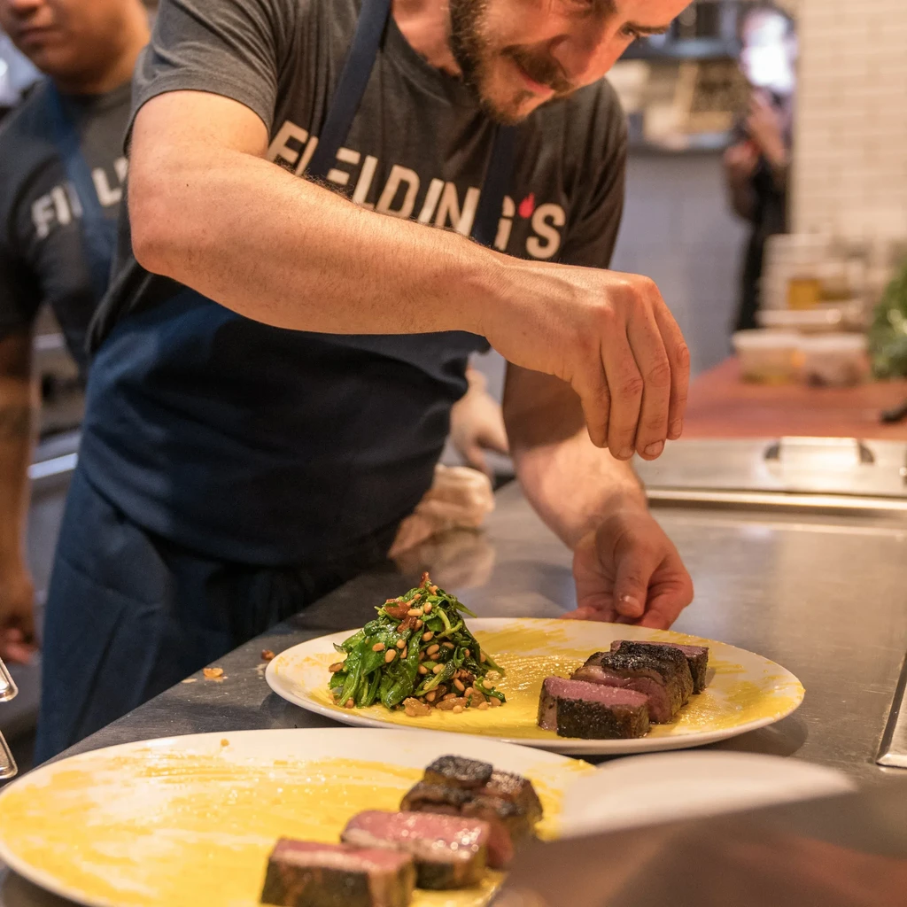 Chef plating seared meat, vegetables, and sauce in a professional kitchen.
