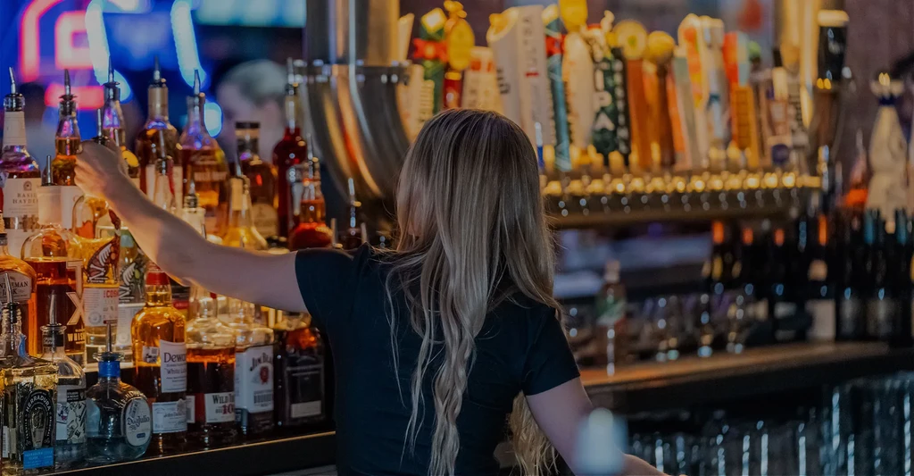 Bartender with long blonde hair reaching for a bottle behind a bar.