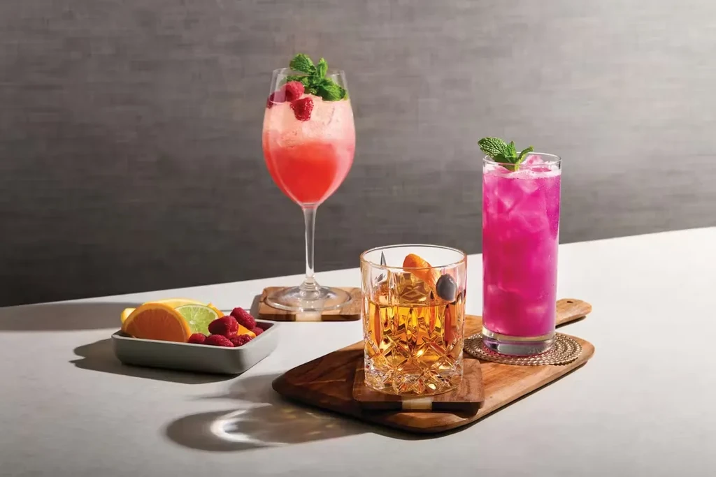Three colorful garnished beverages on a wooden board with fruit.