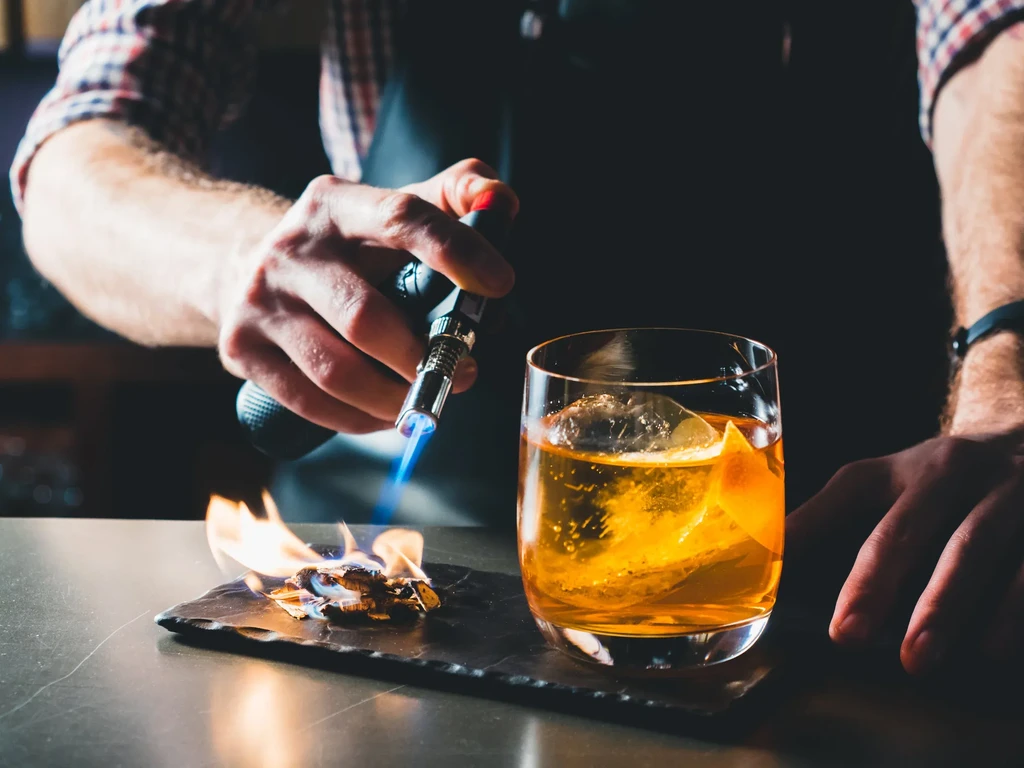 Bartender's hands torching wood chips next to an amber cocktail.