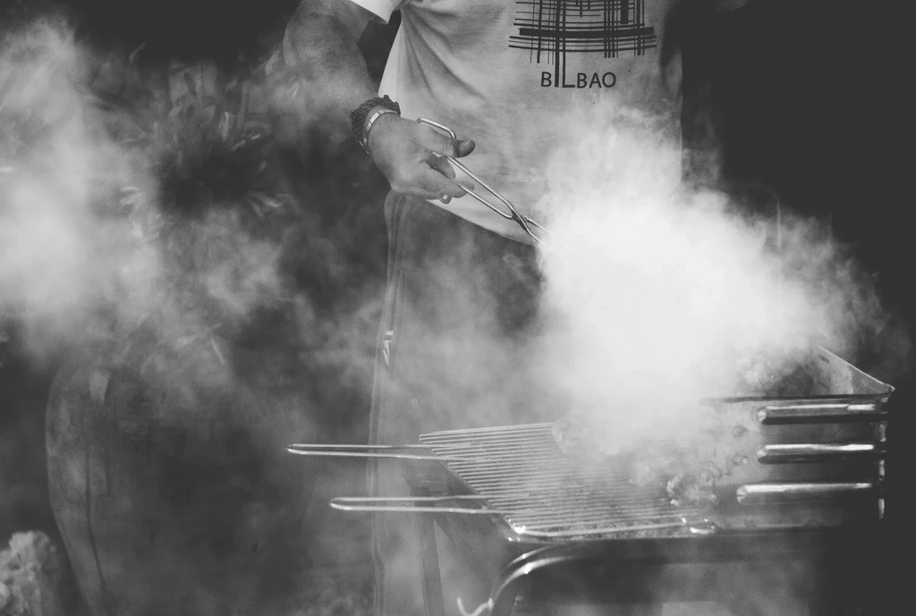 Person grilling food on an outdoor grill, dense smoke rising.