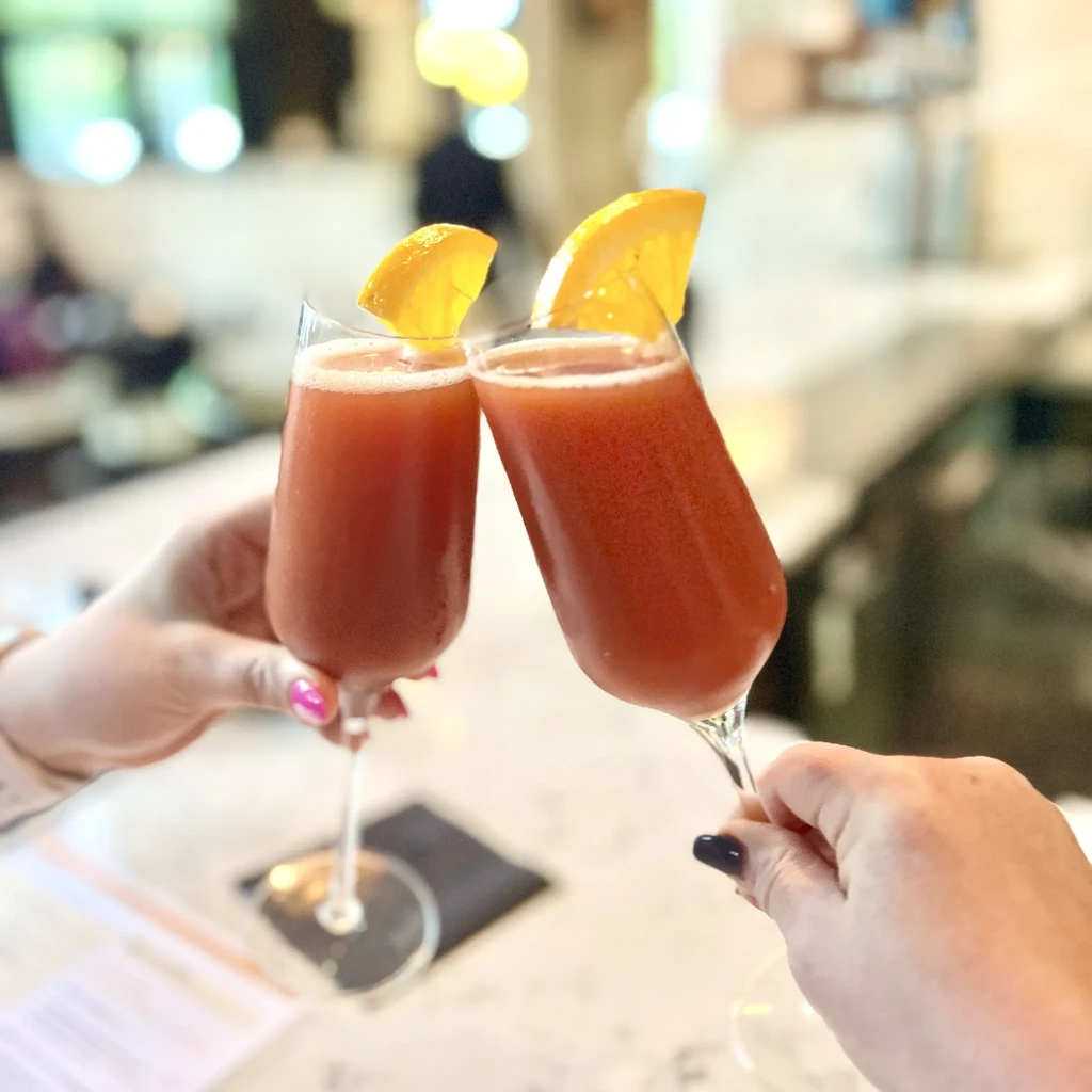 Hands clinking champagne flutes filled with orange cocktails and wedges.