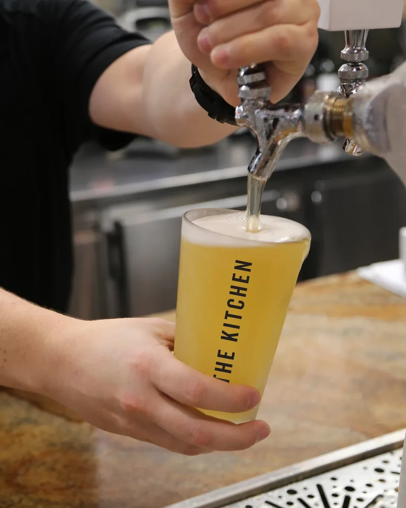 Hands pour frothy beer from a chrome tap into "THE KITCHEN" glass.