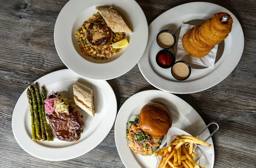 Overhead view of four distinct dishes: burger, onion rings, glazed meat, salad.
