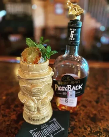 Tiki cocktail in a pig mug, whiskey bottle, and coaster on a bar.
