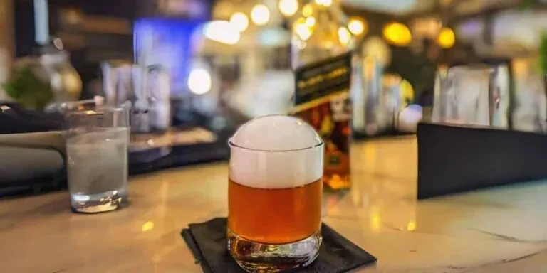 Amber cocktail with a domed white foam on a bar counter.