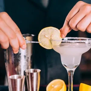 Hands garnishing a margarita with a lemon slice at a bar.