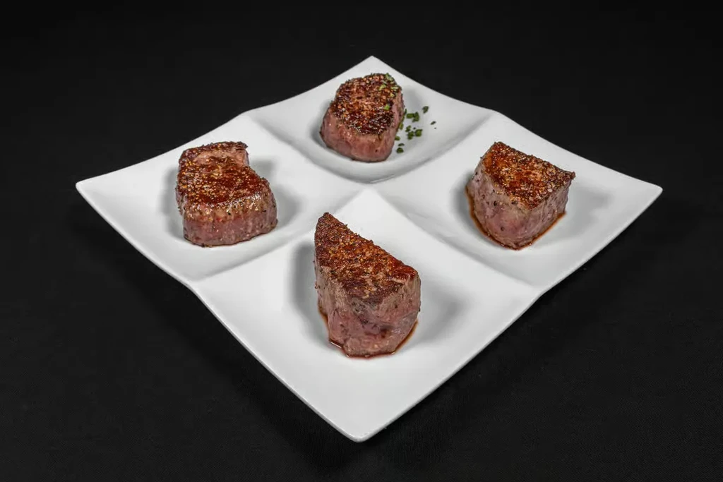 Four seared medium-rare meat pieces with chives on a white plate.