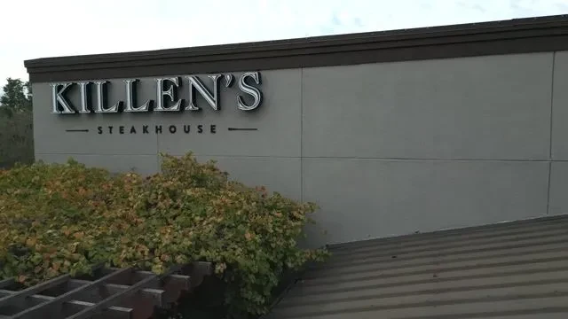 Exterior view of Killen's Steakhouse sign on a gray wall with foliage.