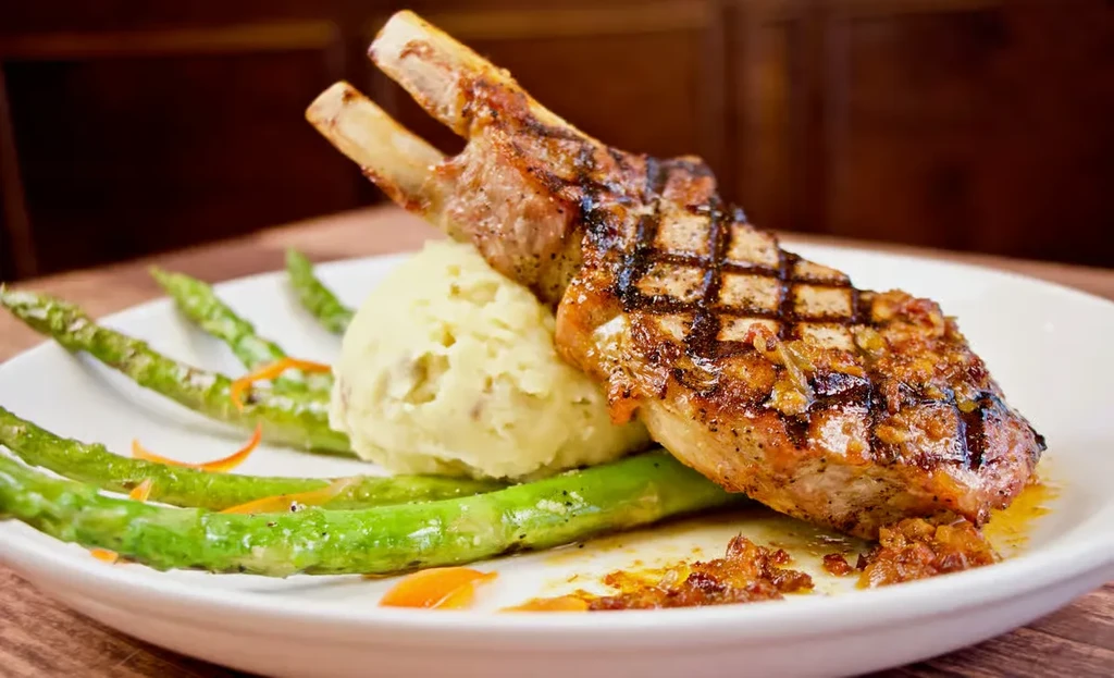 Plated grilled bone-in chop, mashed potatoes, asparagus, orange garnish.