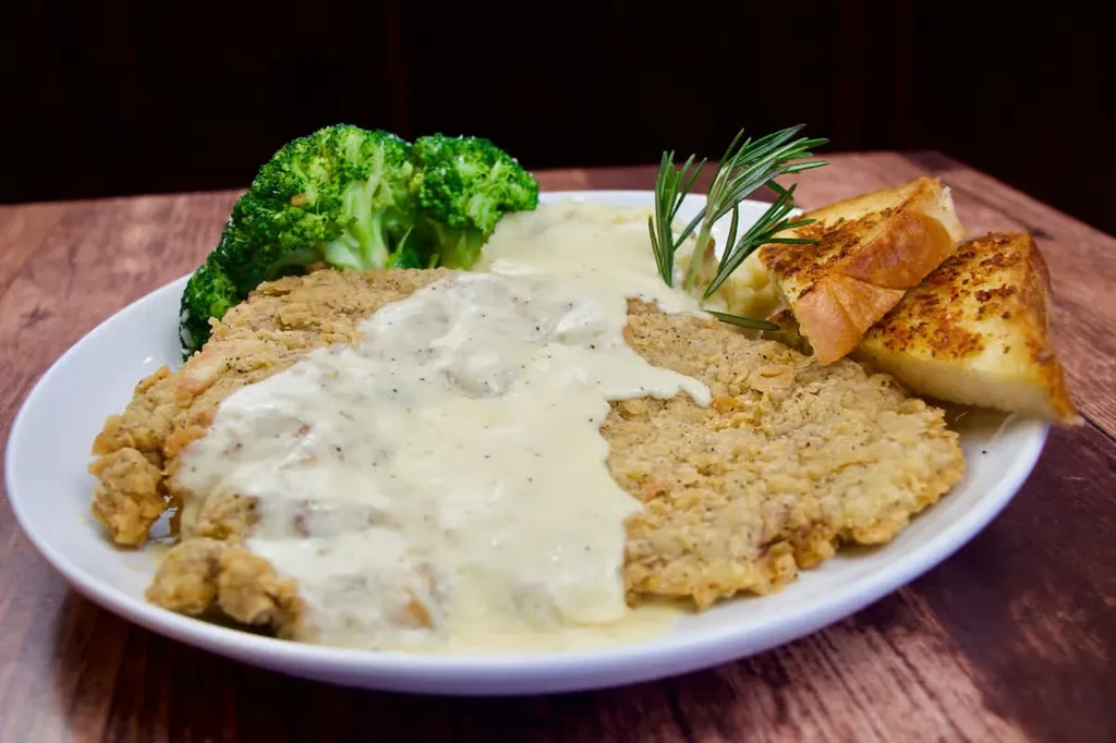 Close-up of fried meat with gravy, broccoli, mashed potatoes, and garlic bread.