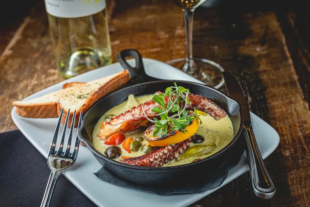 Cast-iron pan with grilled octopus, creamy sauce, vegetables, and bread.