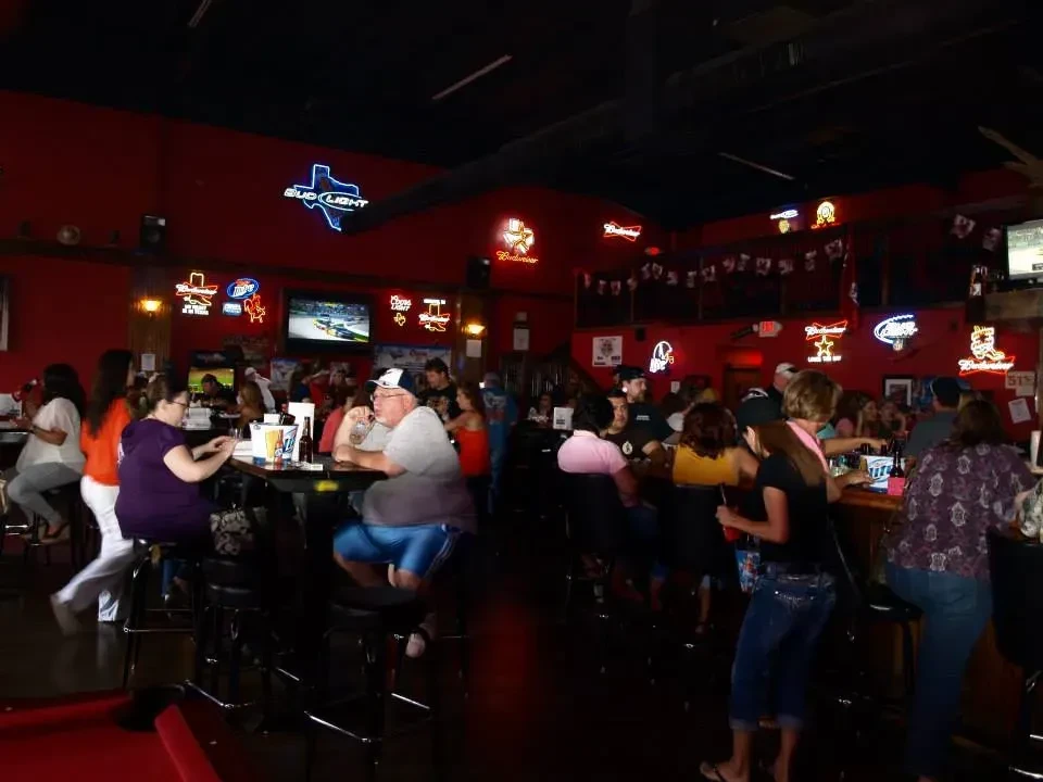 Dimly lit bar with red walls, neon beer signs, and patrons.