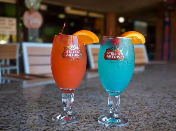 Two colorful cocktails in Stella Artois glasses with orange and cherry garnishes.