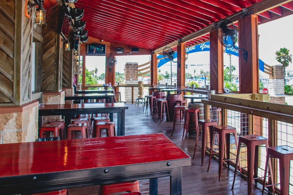 Outdoor restaurant seating area with red tables, dark wood, and boats.