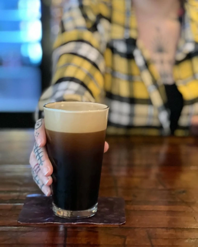 Tattooed hand holds a dark stout with creamy head on a wooden table.