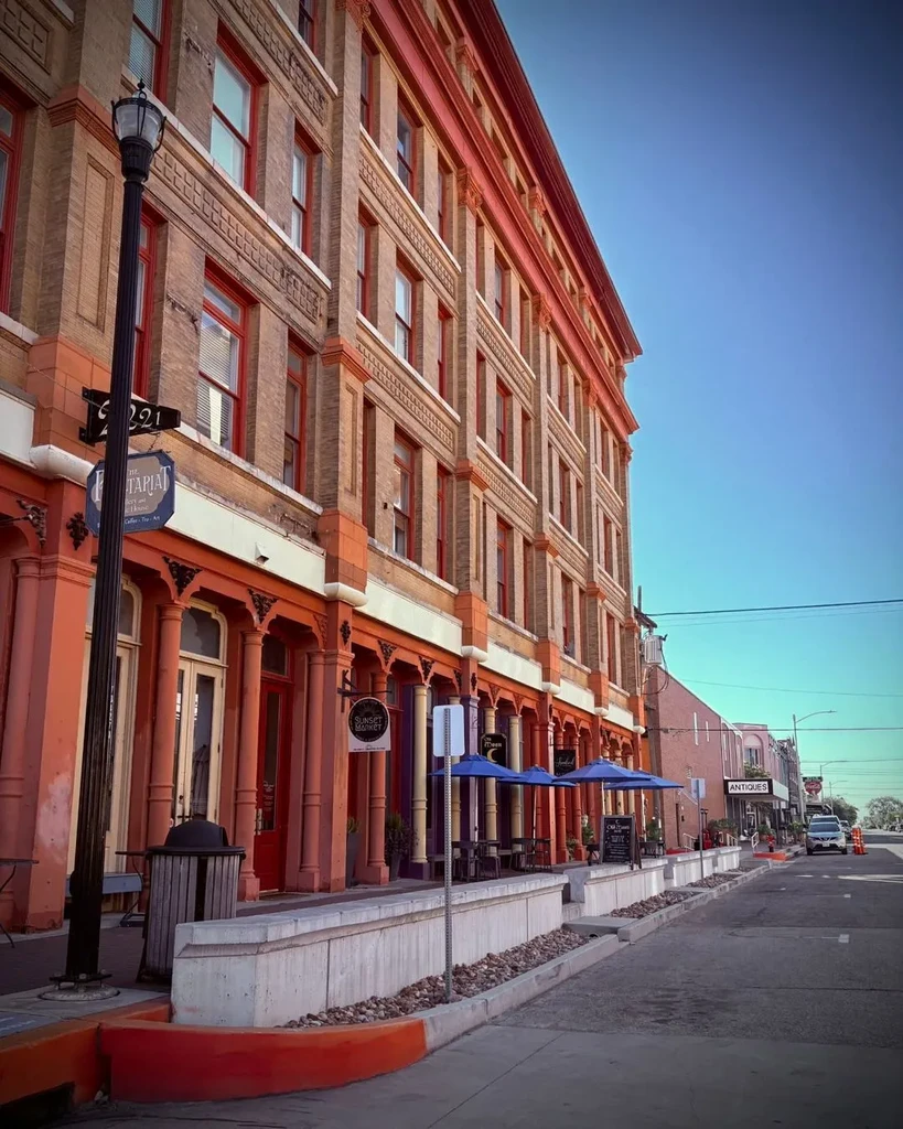 Multi-story brick buildings with red trim and cafes on a sunny street.