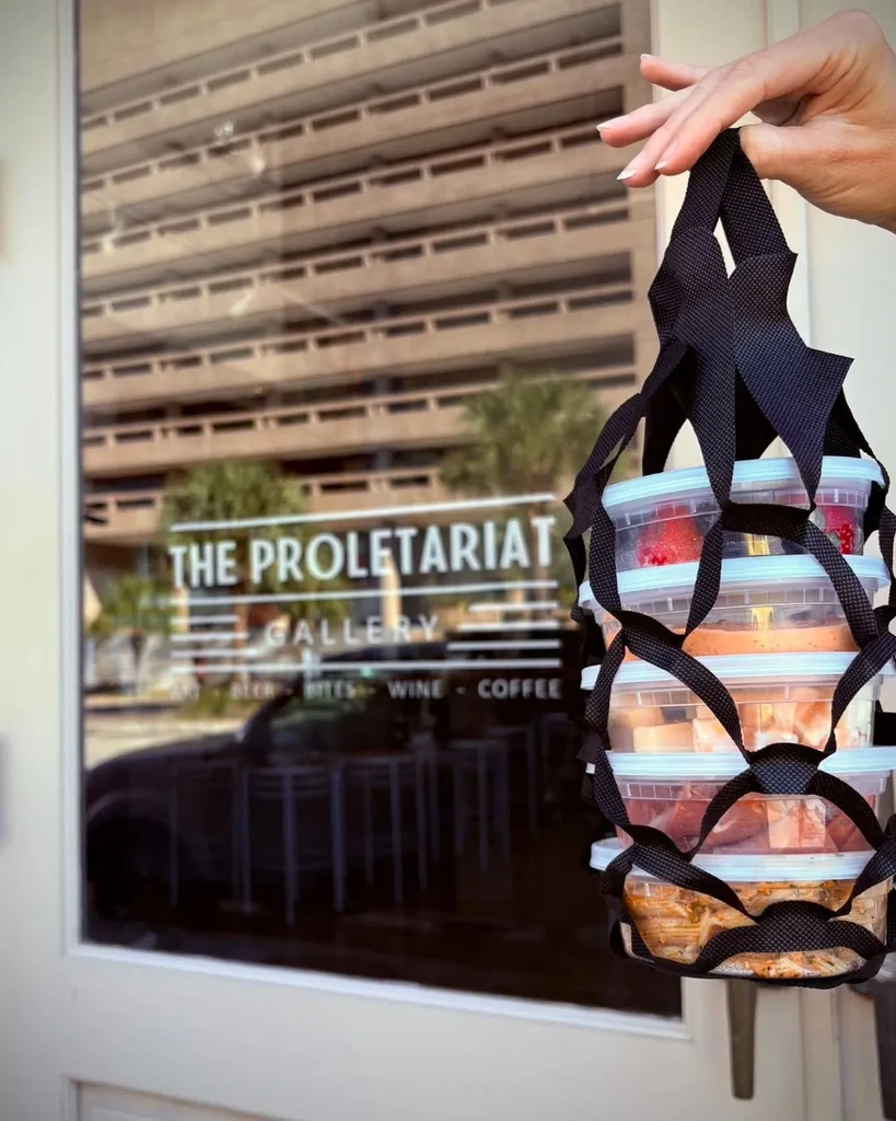 Hand holds mesh bag of takeaway food; "THE PROLETARIAT GALLERY" reflected.