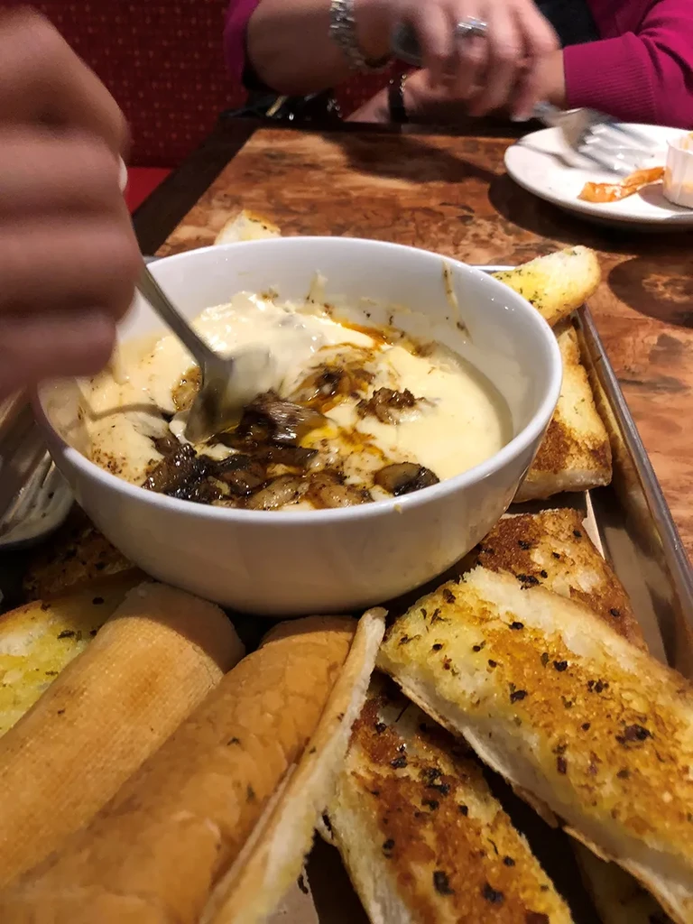 Hand stirring creamy dip with caramelized onions, surrounded by toasted bread.
