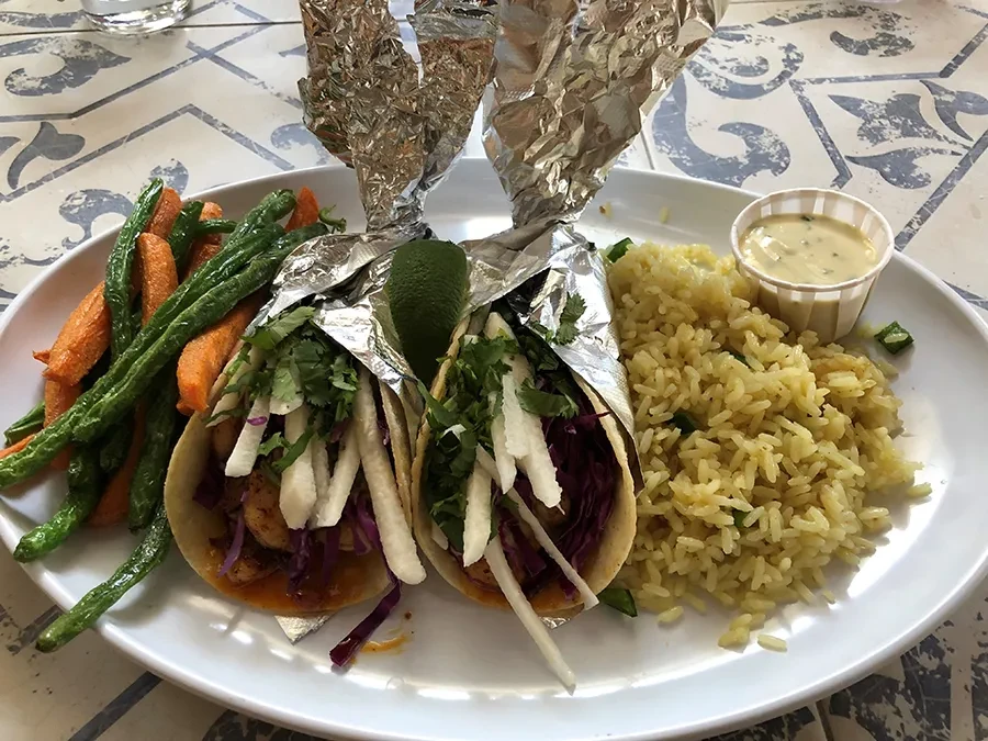 Two foil-wrapped tacos, rice, vegetables, and sauce on a patterned table.