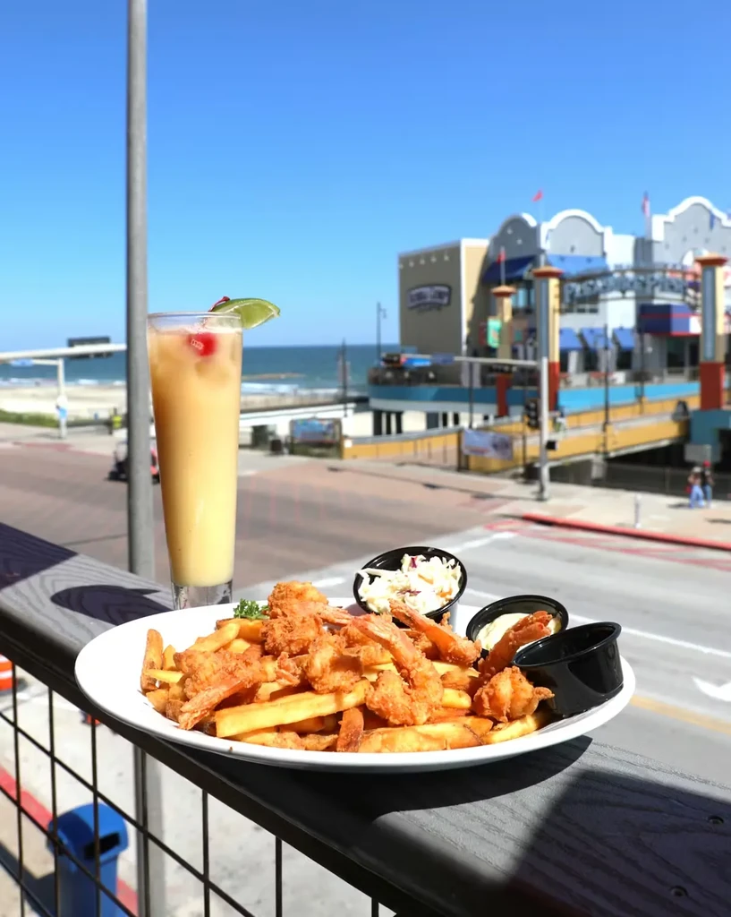 Fried shrimp and fries with a drink by a sunny beach and pier.