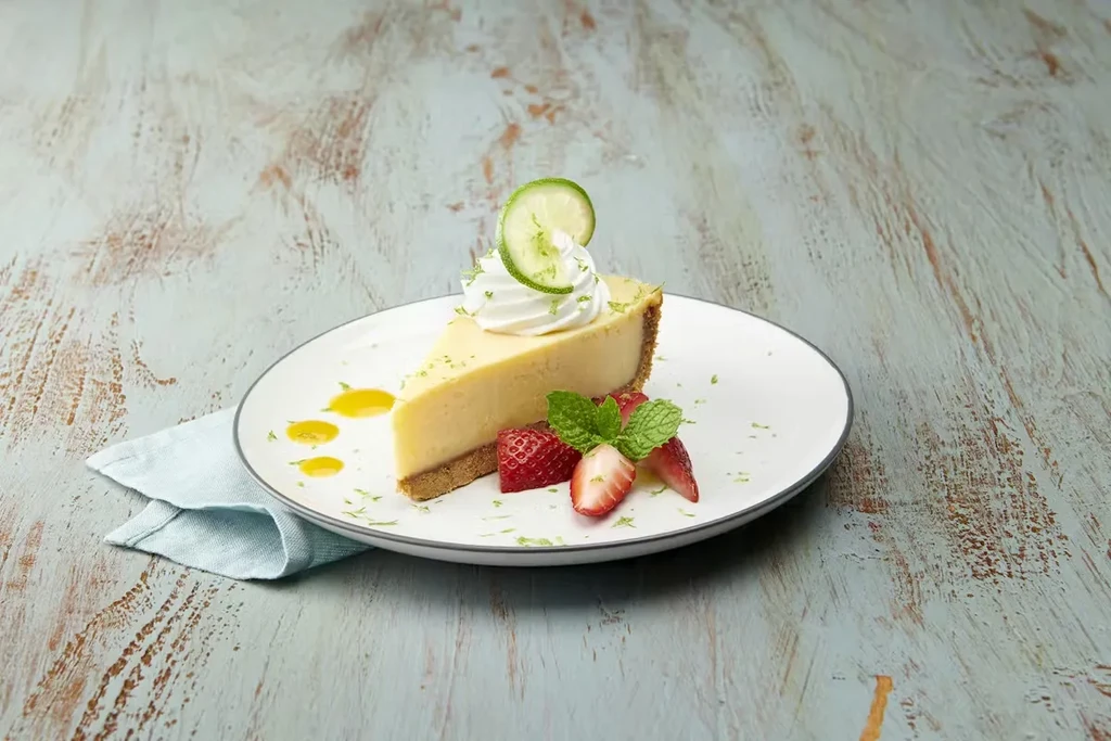 Key lime cheesecake with whipped cream, lime, and strawberries on wood.