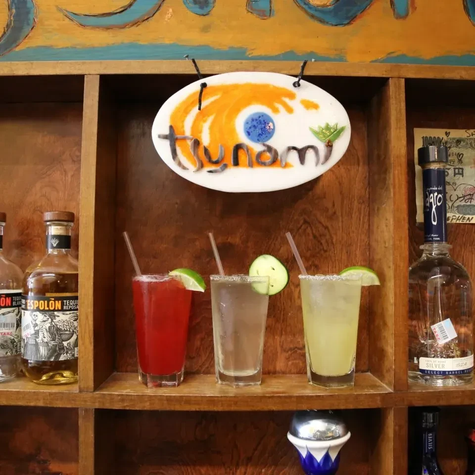 Garnished drinks, tequila bottles, and 'tsunami' sign on rustic shelf.