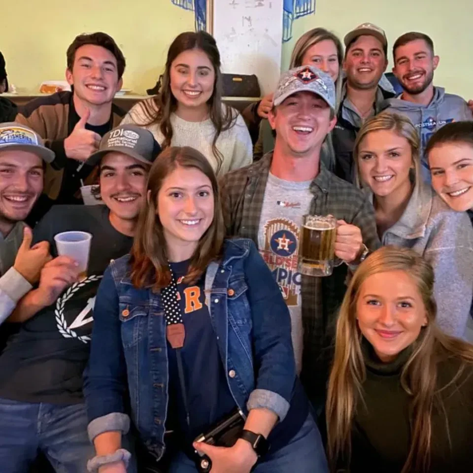 Smiling group of ten young adults holding drinks, casually dressed indoors.
