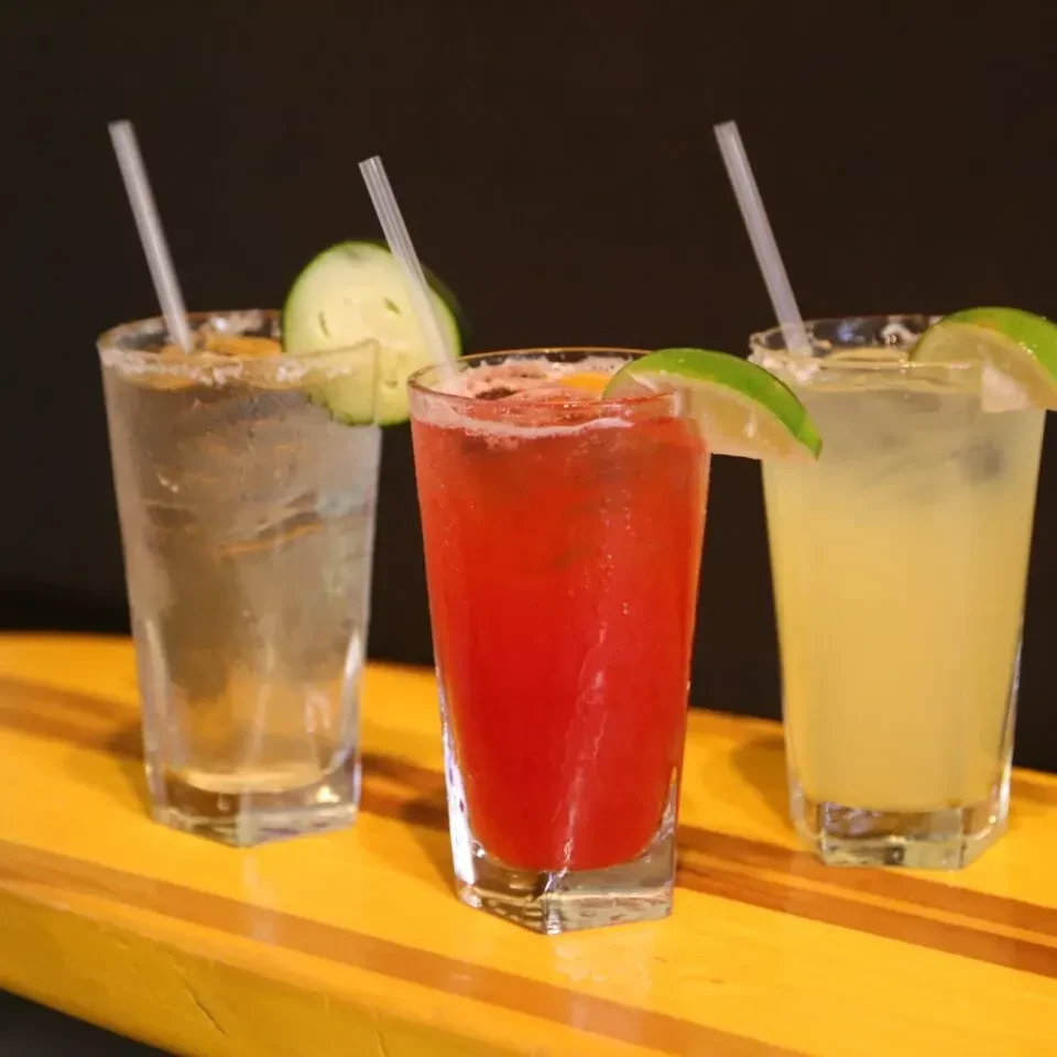 Three tall glasses with colorful beverages, garnishes, straws on yellow.