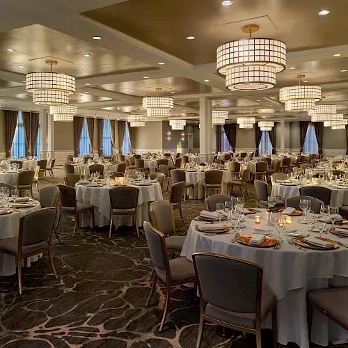 Elegant banquet hall with set round tables, chandeliers, and draped windows.