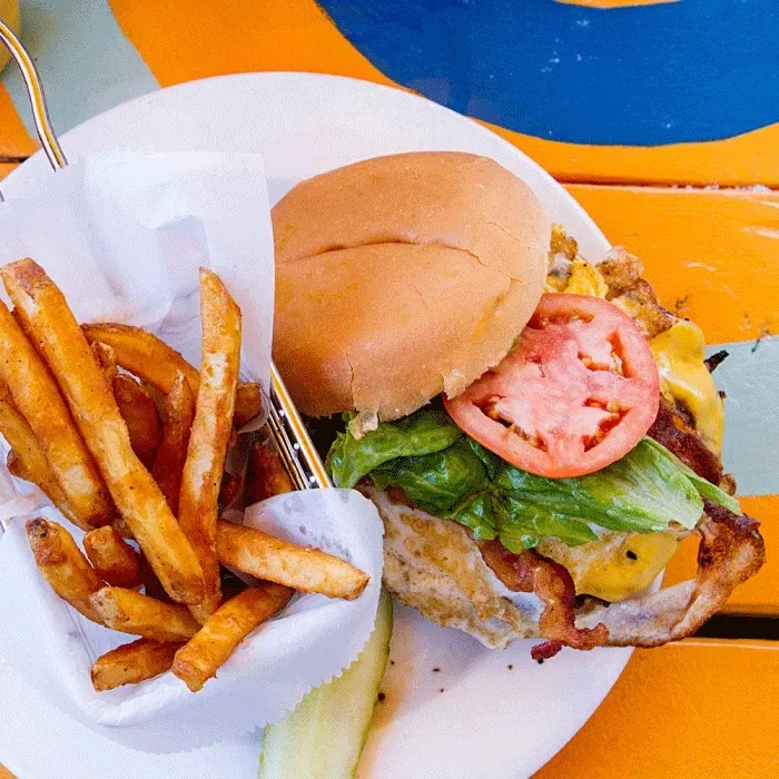 Burger with egg, bacon, fries, and pickle on a vibrant table.