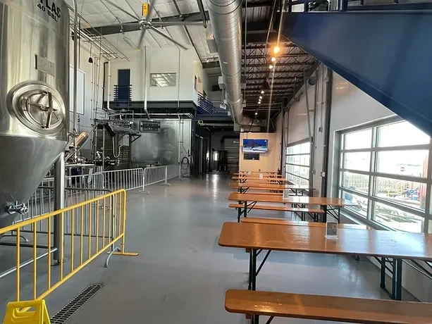 Bright industrial brewery interior with tank, wooden tables, and glass doors.