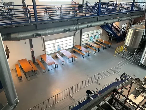 Open-plan industrial brewery with wooden tables, brewing tanks, and mezzanine seating.