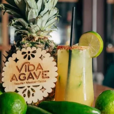 Light yellow-green drink with lime, tajín, pineapple, and "Vida Agave" sign.