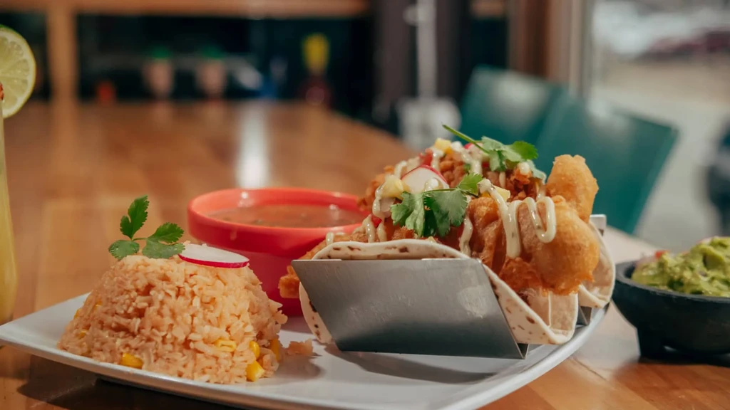 Three tacos, rice, guacamole, and soup served on a wooden table.