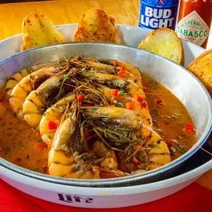 Head-on shrimp in reddish sauce with bread in a metal pan.