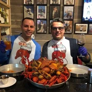 Two men in crawfish bibs smile at a large crawfish boil.