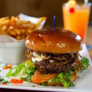 Tall gourmet burger, golden fries, and a blurred orange drink.