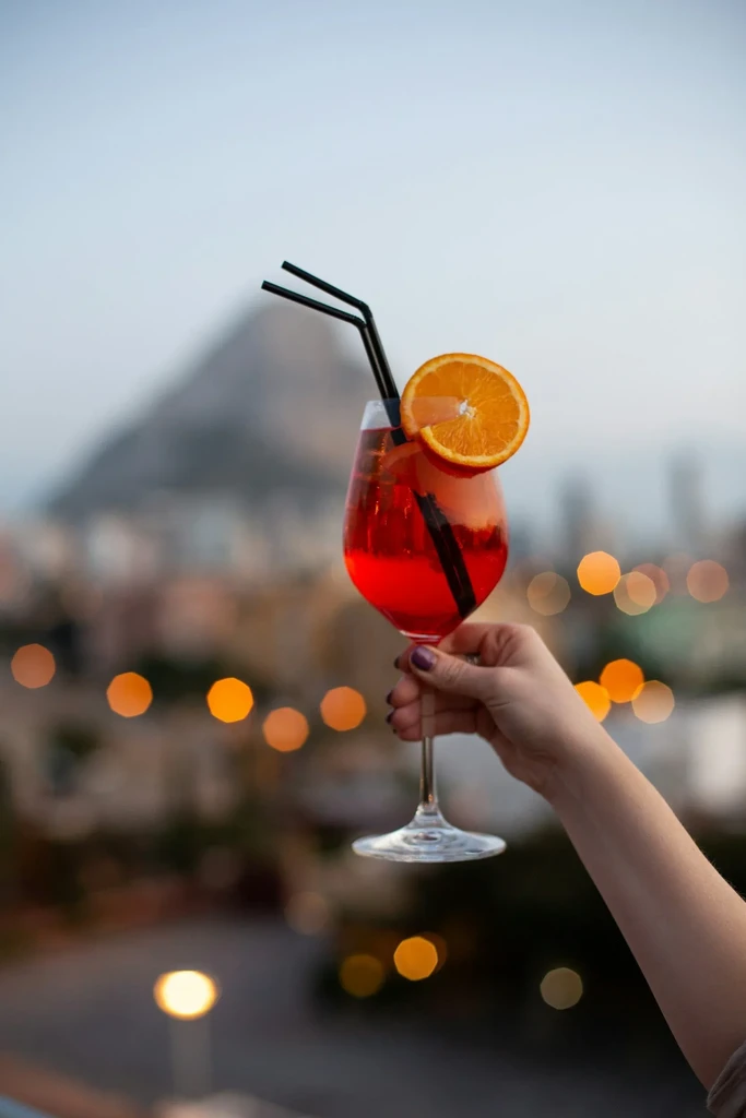 Hand holding red-orange cocktail with orange, straws, against blurred city lights.