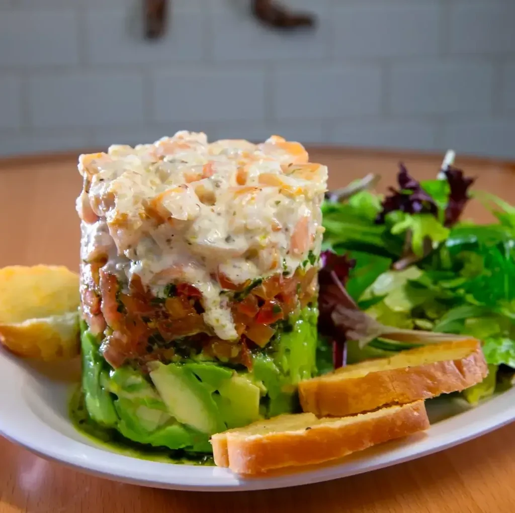 Vertically layered avocado, tomato, shrimp dish with side salad and bread.