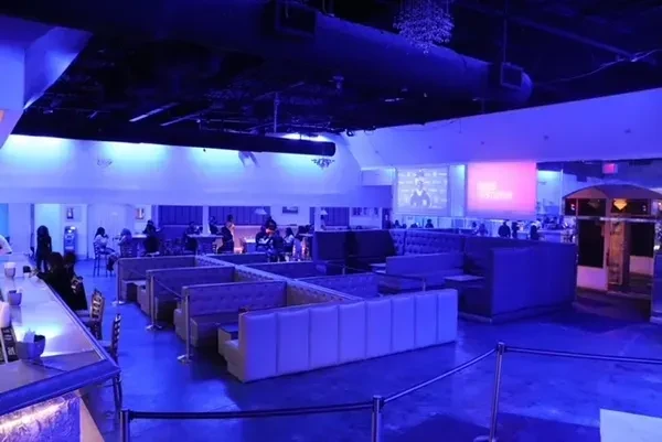 Blue-lit modern lounge with bar, booth seating, and two large screens.