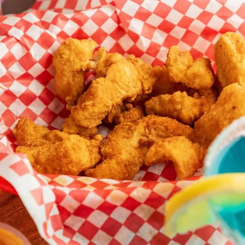 Crispy golden-brown fried food in a checkered basket; blurred drink visible.