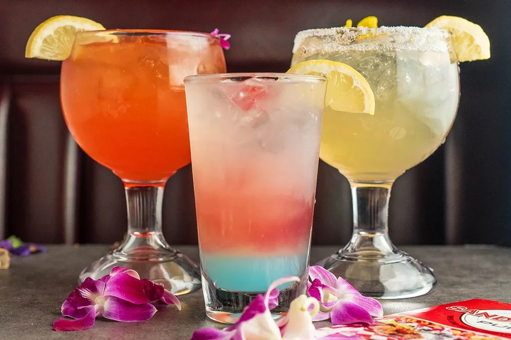 Three colorful cocktails, garnished with lemon and purple orchid petals.